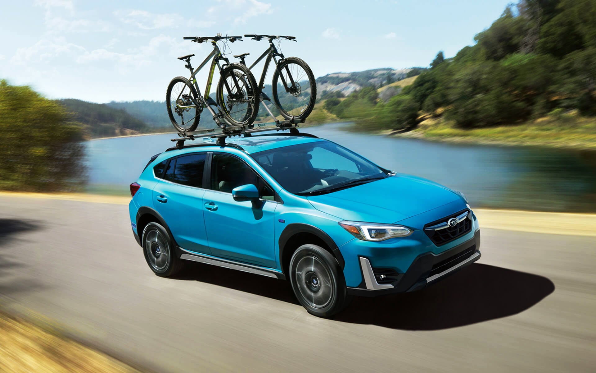 A blue Crosstrek Hybrid with two bicycles on its roof rack driving beside a river | Romano Subaru in Syracuse NY